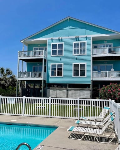 Turquoise beach house on stilts with white railings, swimming pool, lounge chairs, and palm trees under clear blue sky