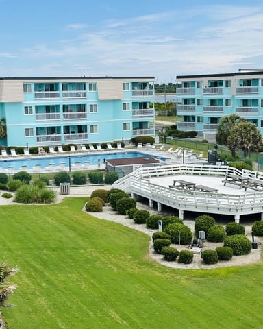 Turquoise beachfront resort with multi-story buildings, swimming pool, white fire pit structure, and manicured lawn with palm trees.