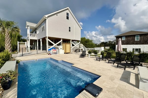 Elevated white house with pool, patio, lounge chairs, and neighboring brown cottage in coastal setting