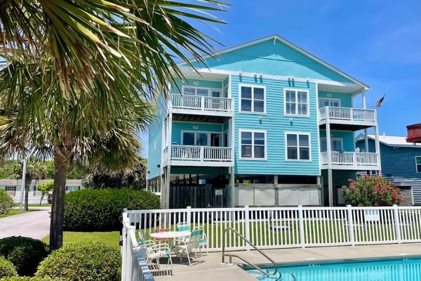 Turquoise beach house with white railings, multiple decks, and swimming pool in foreground under clear blue sky