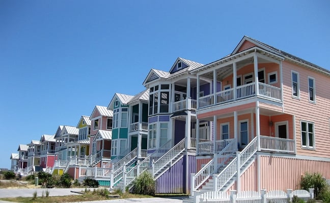 Row of colorful elevated beach houses with white railings and metal roofs along sandy shore under clear blue sky