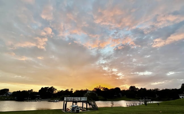 Waterfront property at sunset with dock, green lawn, and dramatic orange and pink sky over calm water